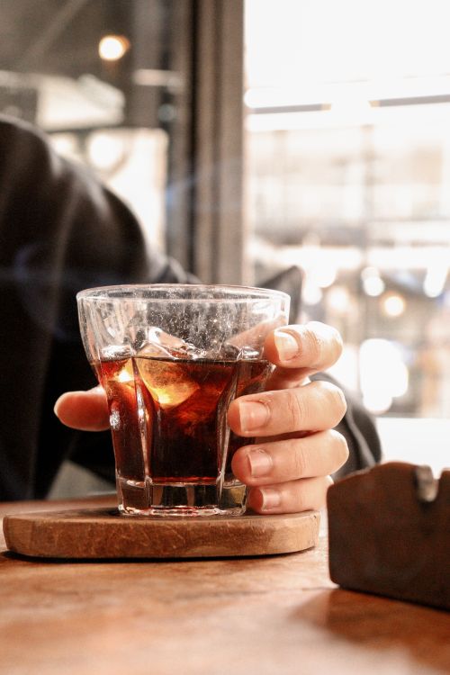 Free A hand holding a glass of iced beverage on a wooden table in a cozy indoor setting. Stock Photo