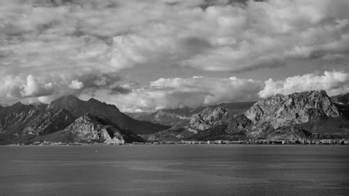 A Grayscale Photo of Mountains Near the Body of Water Under the Cloudy Sky