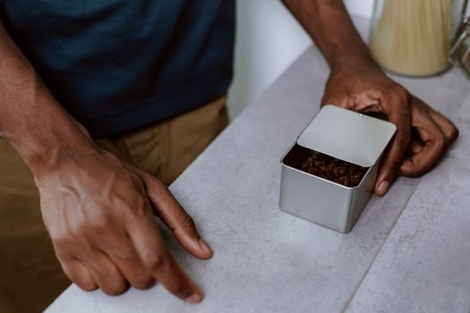 A man holds a metal container filled with fresh coffee beans on a kitchen counter.