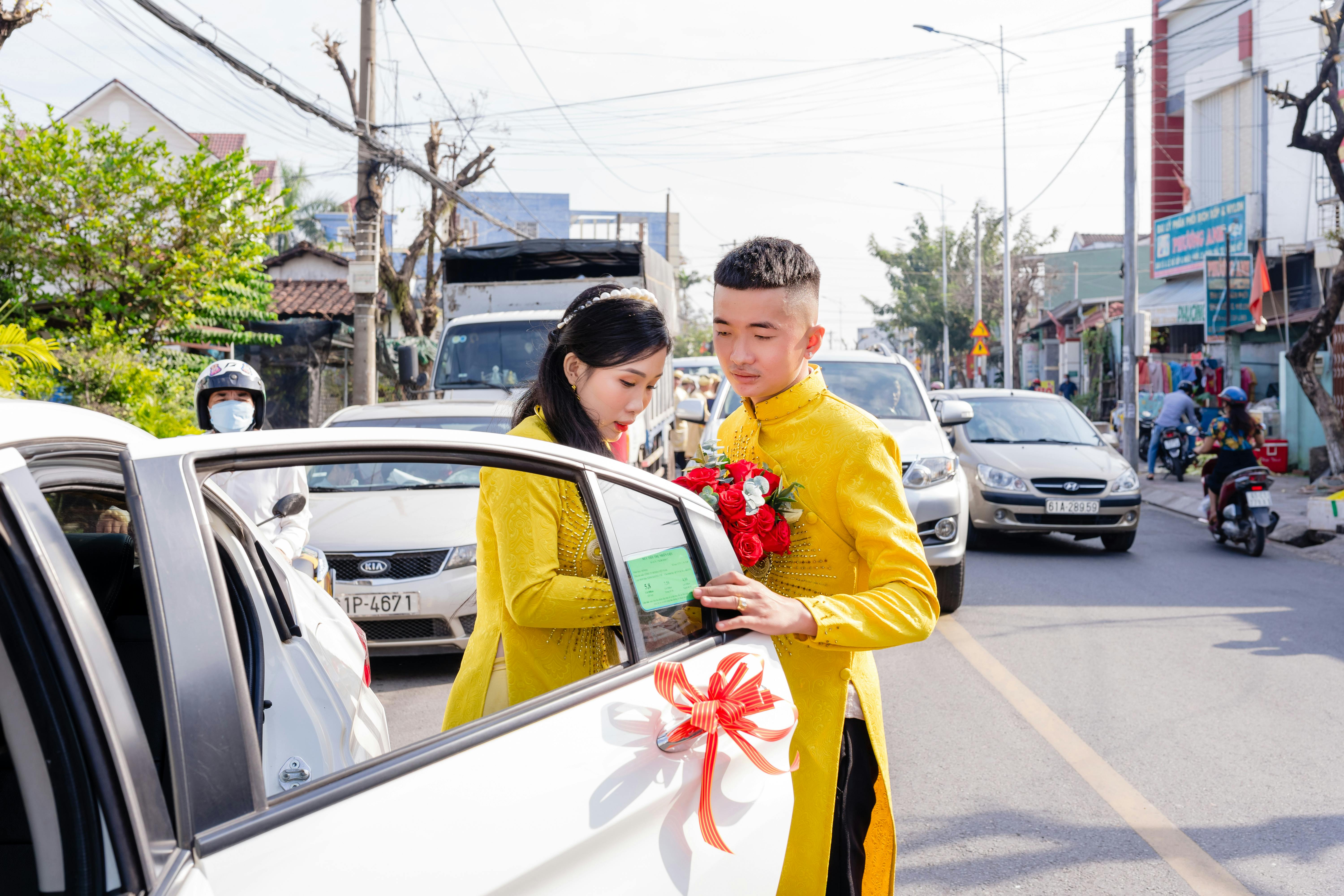 Young Vietnamese couple in traditional wedding outfits exiting a decorated car on a busy street.