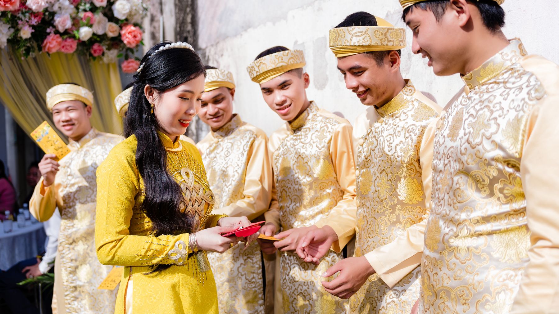 Group of people wearing traditional Vietnamese ao dai during a wedding ceremony, exchanging red envelopes.
