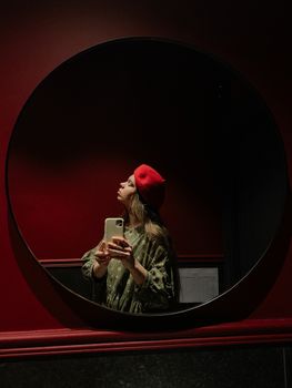 A woman wearing a red beret takes a selfie in a round mirror against a dark red background.