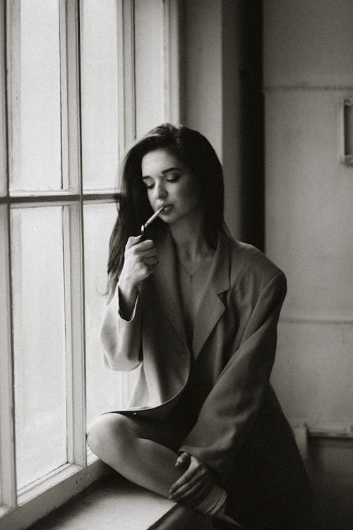 A Grayscale Photo of a Woman Smoking Cigarette while Sitting Near the Window