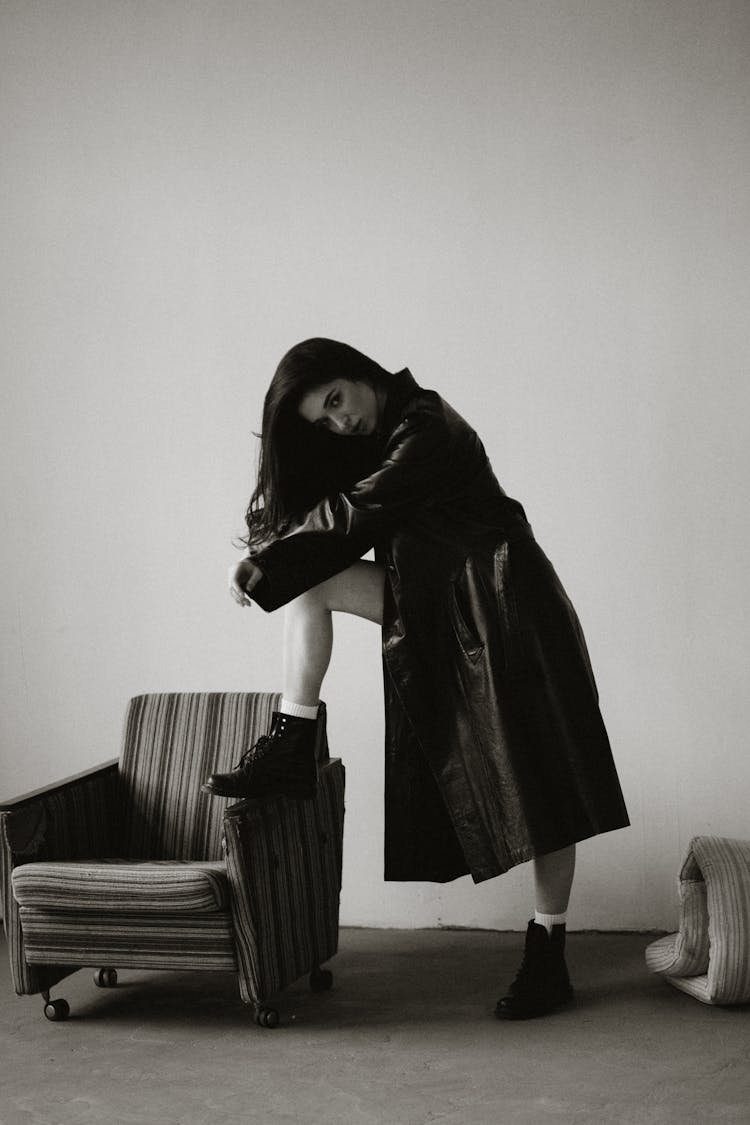 Woman With Long Black Hair In Leather Coat Standing With One Leg On Armchair