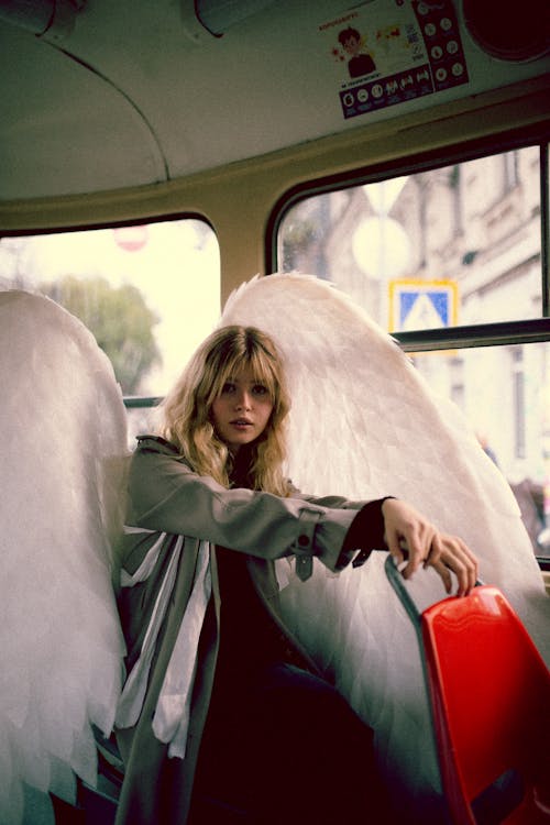 Blond Woman in Coat and with Angel Wings Sitting in City Bus