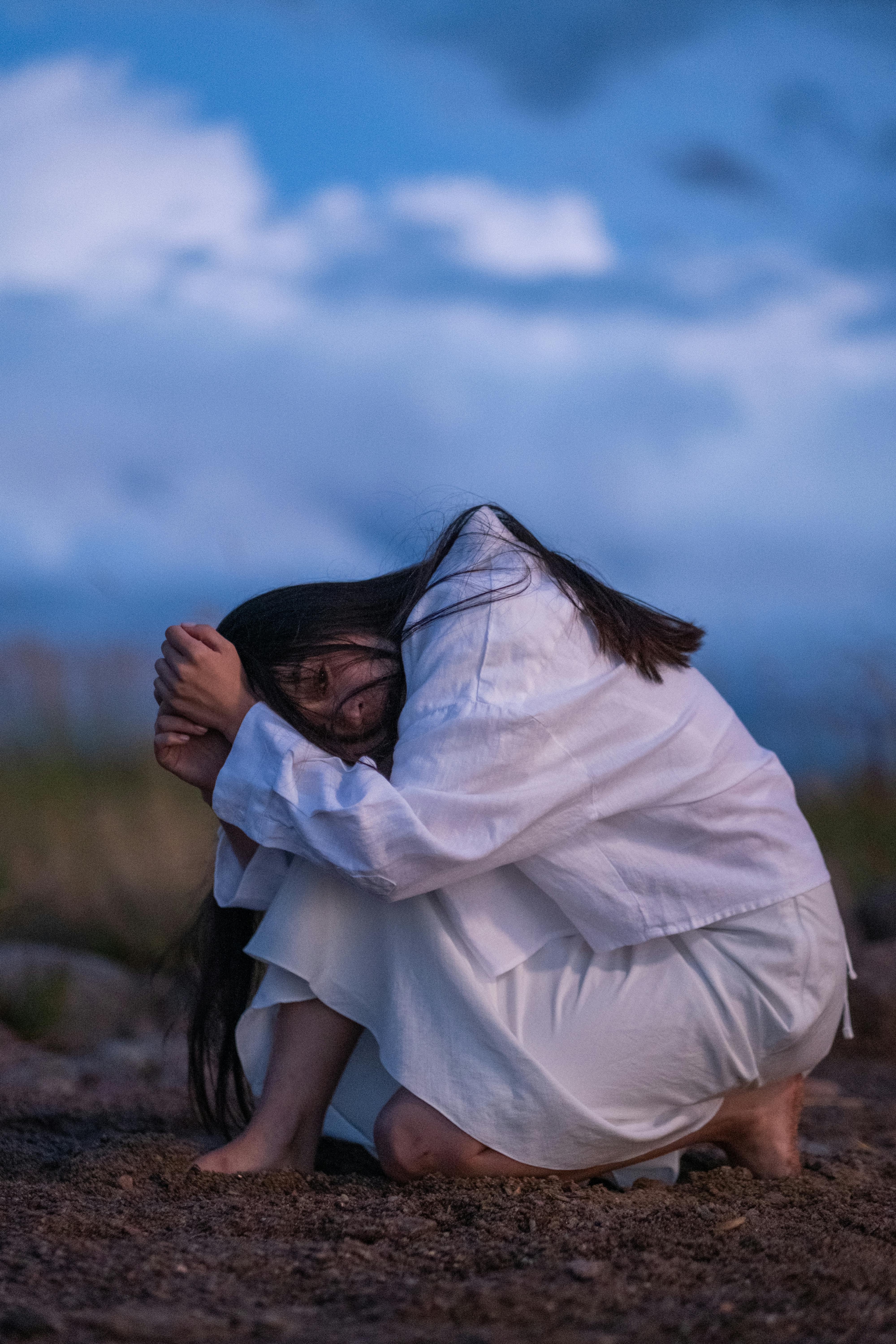 Woman in a white dress crouching outdoors, enveloped in thoughtful contemplation.