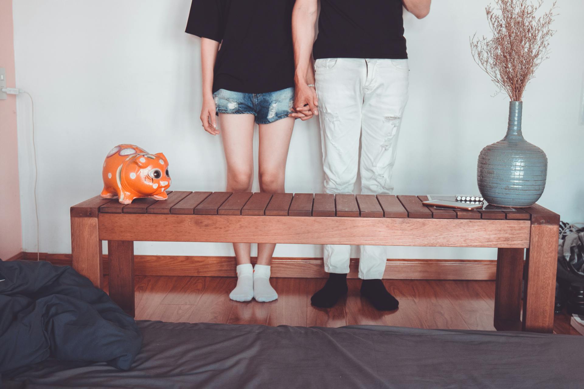 A couple holds hands inside a cozy room, featuring a piggy bank and a wooden table.