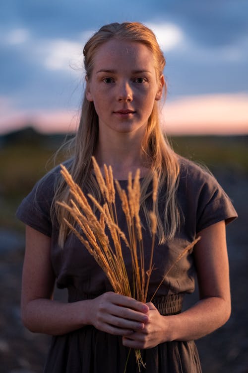 Woman in Brown Crew Neck T-shirt Holding Wheat Flowers while Seriously Looking at Camera