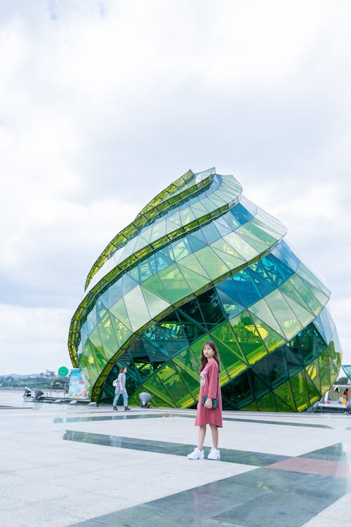 Free Woman in Red Dress Standing in Front of Green Glass Elliptical Building Stock Photo