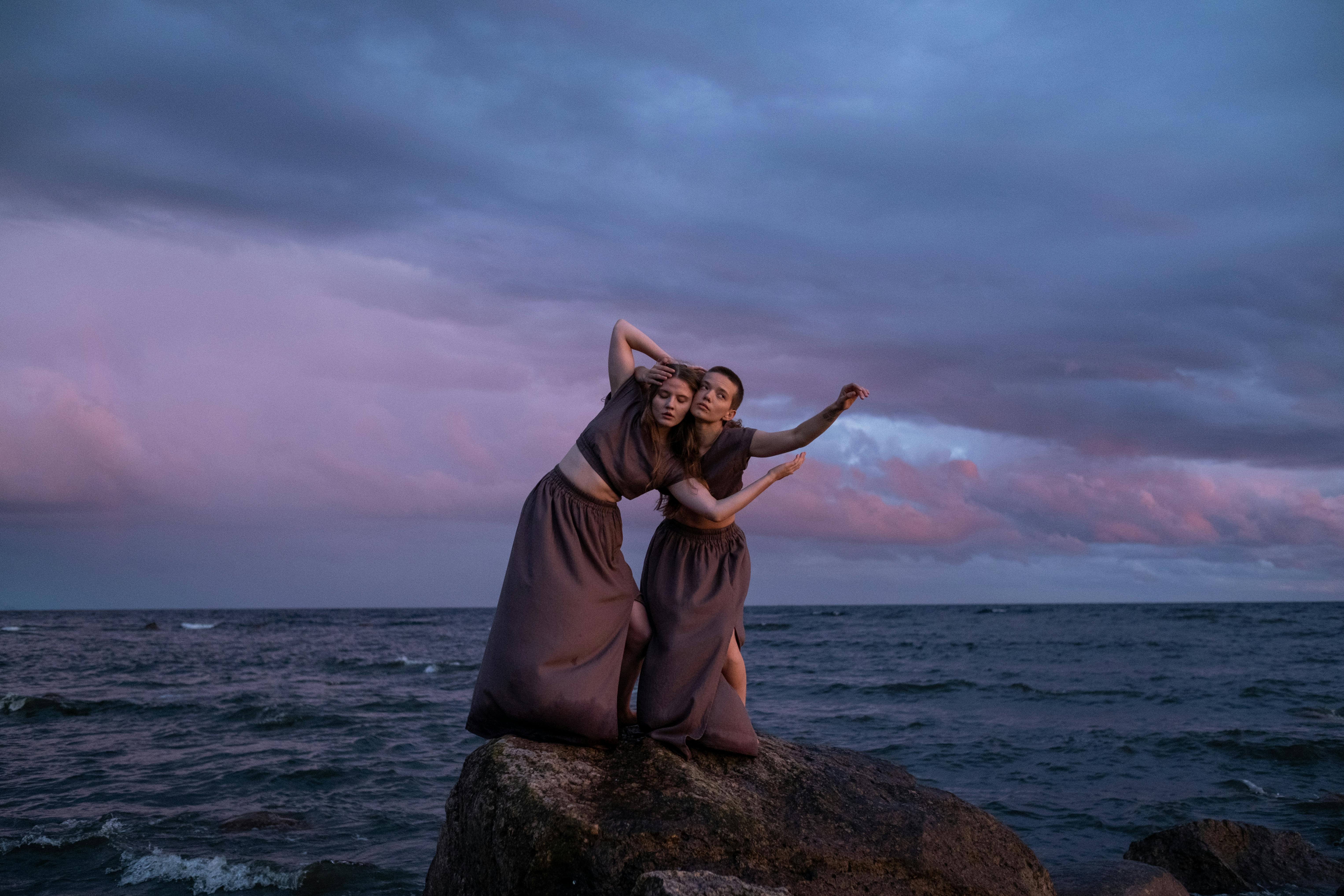 Two dancers pose gracefully on rocks by the sea under a dramatic twilight sky.