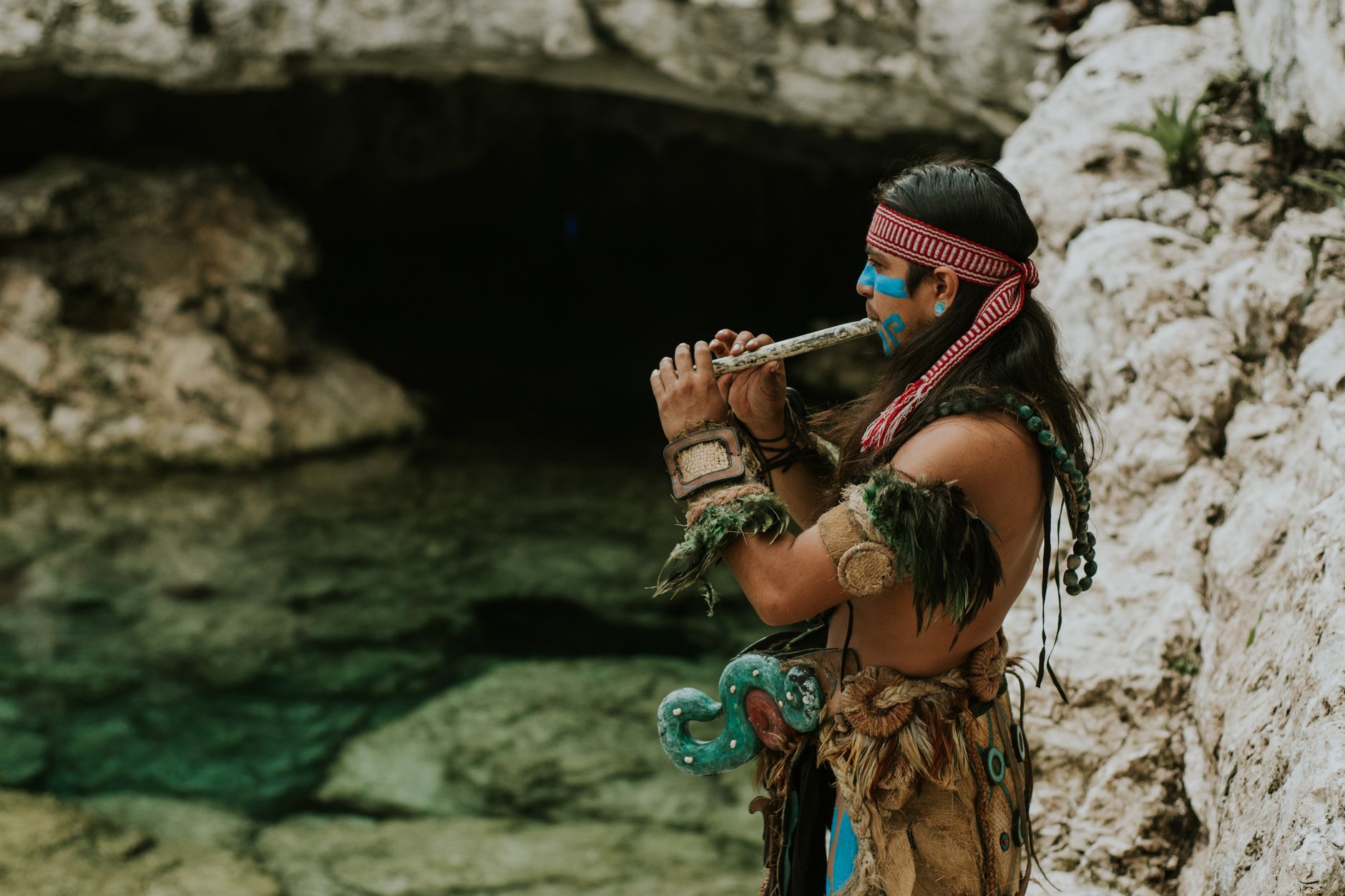 A Mayan musician wearing traditional attire plays a flute by a rocky water landscape, exuding cultural heritage and art.