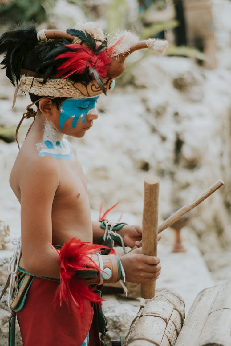 Boy In Traditional Maya Clothes And Face Paint Holding Wooden Drumsticks