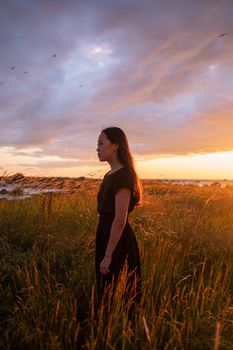 A woman stands amid tall grass, gazing at a stunning sunset near the sea.