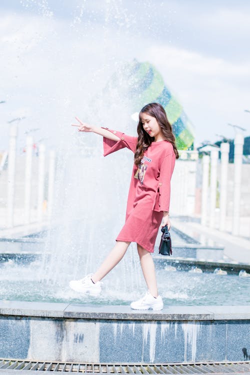 Woman Wearing Pink Long-sleeved Dress Walking on Fountain during Daytinme