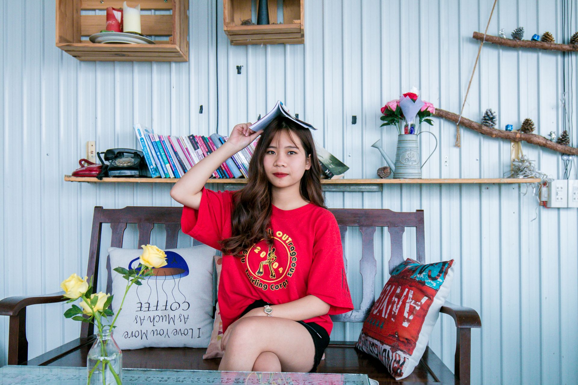 A young woman in a red shirt humorously holds a book on her head while sitting indoors.