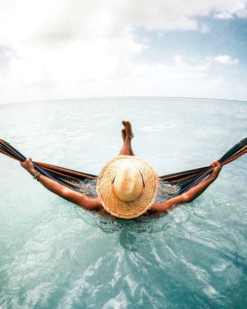 Free A person in a straw hat relaxes in a hammock above calm turquoise waters with a bright sky in the background. Stock Photo