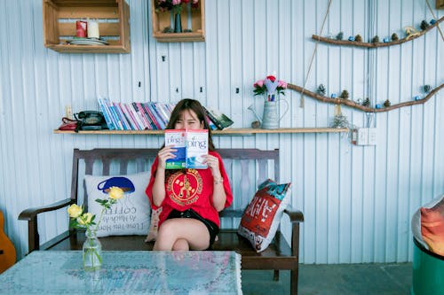 Женщина в красной рубашке держит книгу, сидя на скамейке перед журнальным столиком