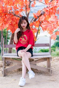 Asian woman sitting on a bench under vibrant orange autumn leaves, enjoying a peaceful moment.
