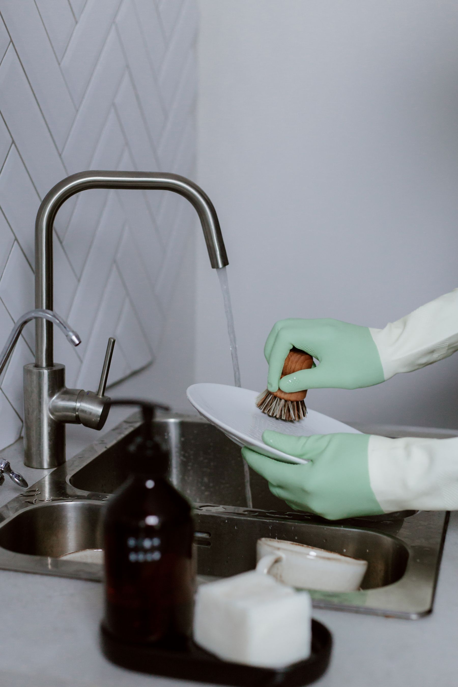 Hands washing a ceramic plate at a kitchen sink with running water and kitchen cleaning tools