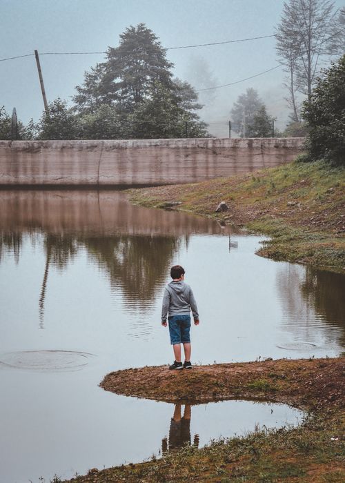 Free A young boy in a hoodie stands by a misty lake, reflecting on nature. Stock Photo