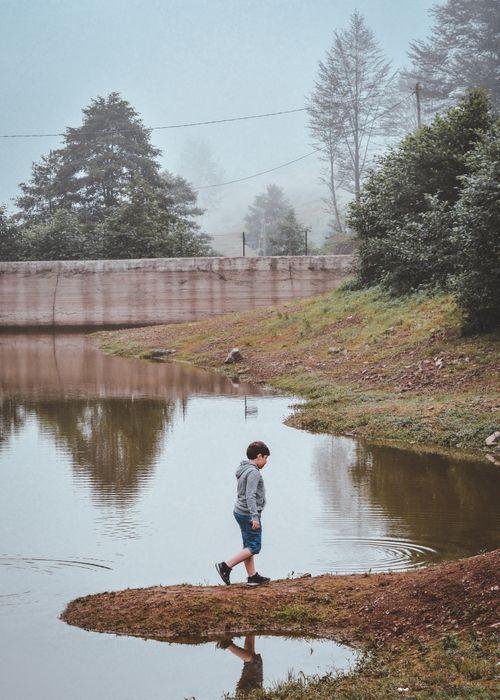 Free A young boy walks along a peaceful lakeside, reflecting trees and foggy atmosphere, nature tranquil moment. Stock Photo
