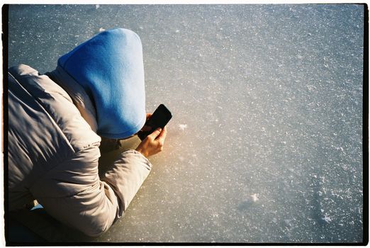 A person in a blue hoodie capturing a photo on a snowy surface with a smartphone.
