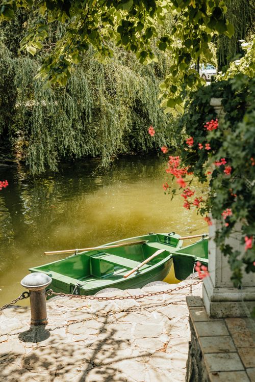 Free A tranquil scene of a green boat moored by a lush, flower-adorned riverside in summer. Stock Photo