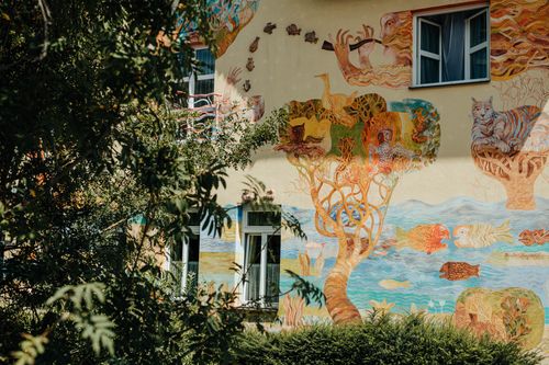 Free Vibrant mural depicting whimsical scenes on a residential building wall surrounded by greenery. Stock Photo