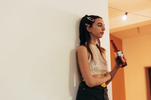 Free Stylish young woman leaning against a wall indoors, holding a beer bottle, looking relaxed. Stock Photo