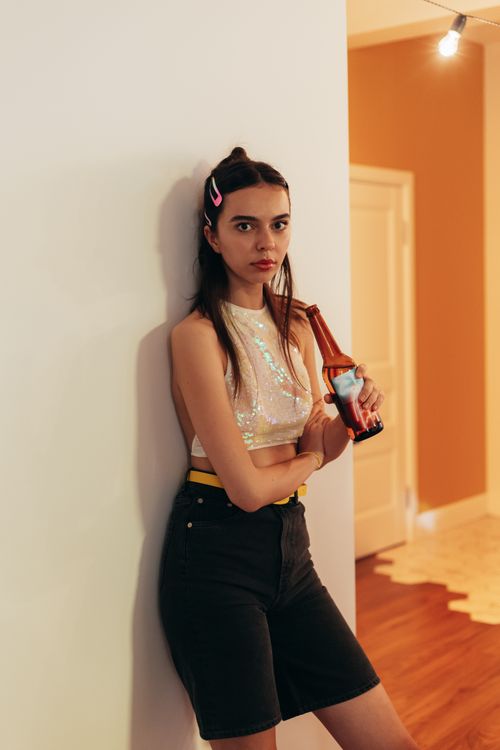 Free Fashionable young woman leaning against a wall, holding a beer bottle in a modern indoor environment. Stock Photo