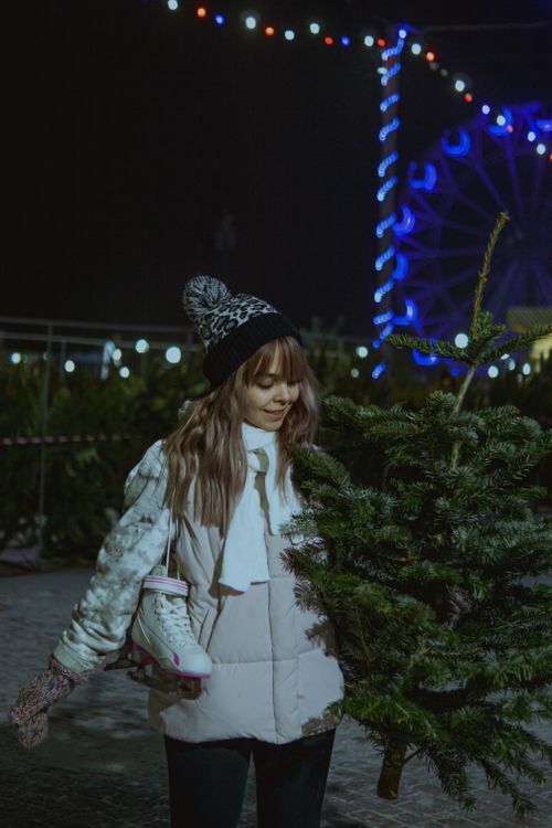 Free Cheerful woman wearing beanie carries pine tree in festive outdoor market with lights. Stock Photo