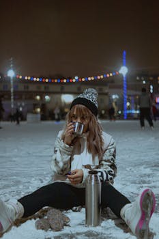 A woman in a beanie drinks from a cup on a snowy night, surrounded by festive lights.