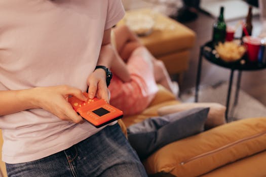 Person playing handheld game on couch, surrounded by relaxed setting with snacks.
