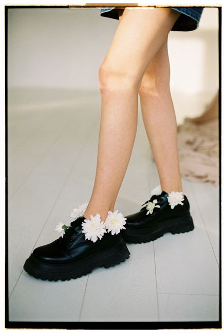 Black leather shoes with white flowers on a light wooden floor, showcasing modern style.