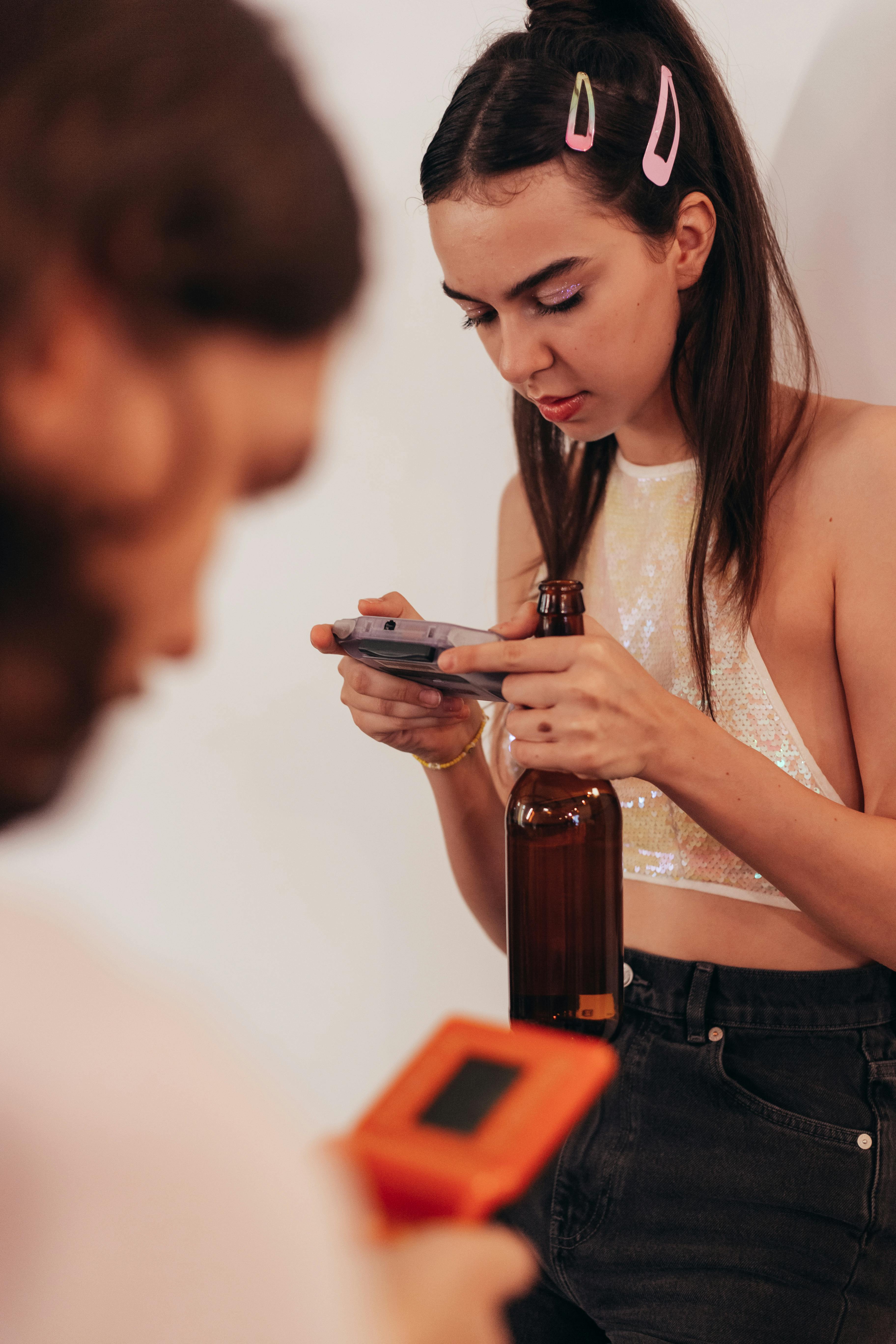 A young woman in a casual setting playing with a retro game device while holding a beer bottle.