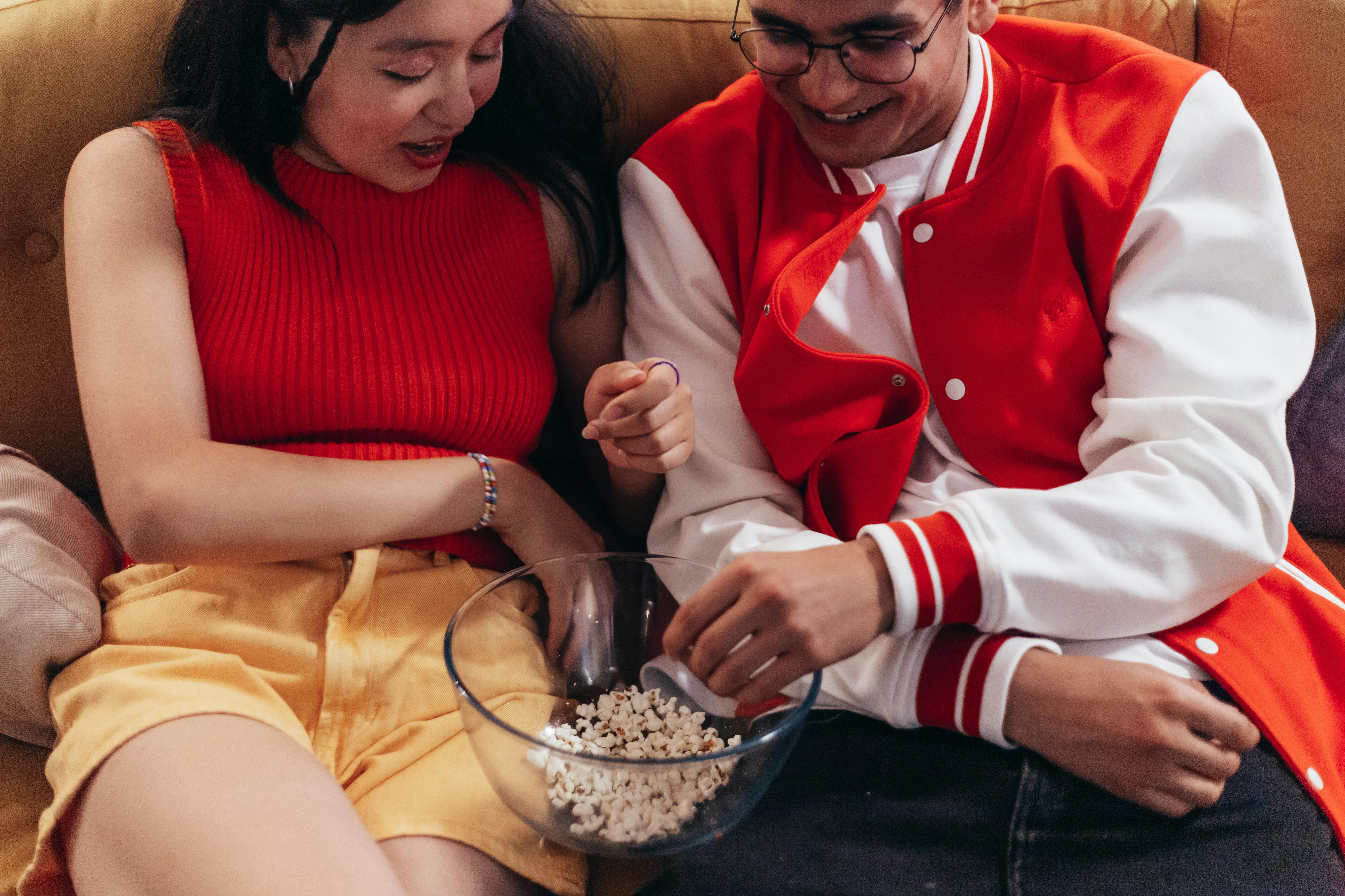 A happy couple relaxing together on a couch, sharing a bowl of popcorn indoors.