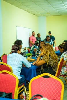 A diverse group of adults in a team meeting, seated and discussing at a table indoors.