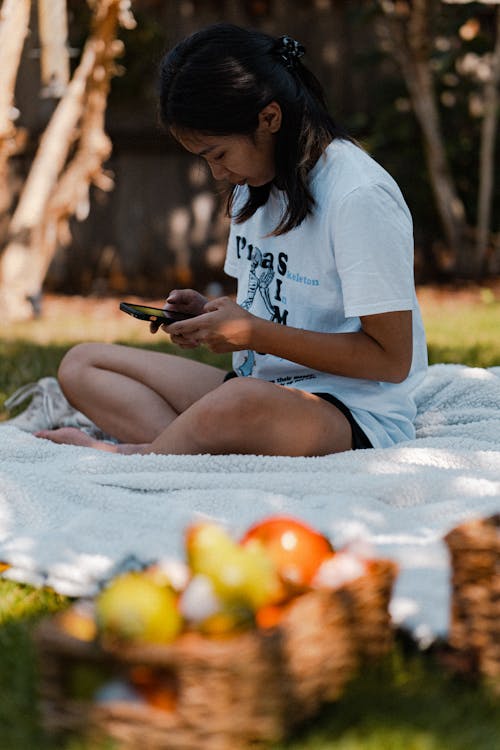 Free Woman in White Shirt Sitting on Blanket While Using Cellphone Stock Photo