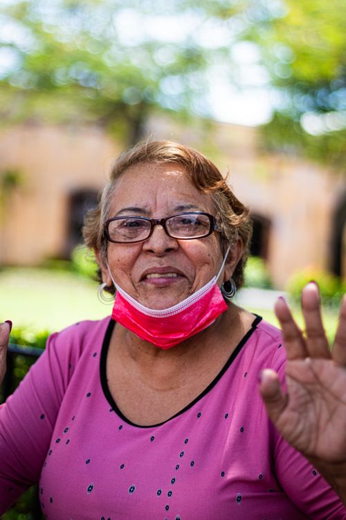 Free Smiling senior woman in pink blouse waves while wearing a face mask outdoors. Stock Photo