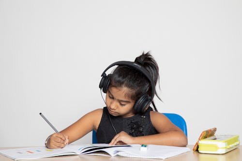 A Girl Wearing Headphones Writing in a Notebook during an Online Class