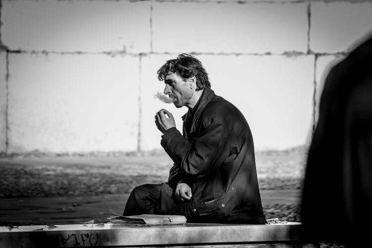 Grayscale Photo Of Smoking Man While Sitting On Bench