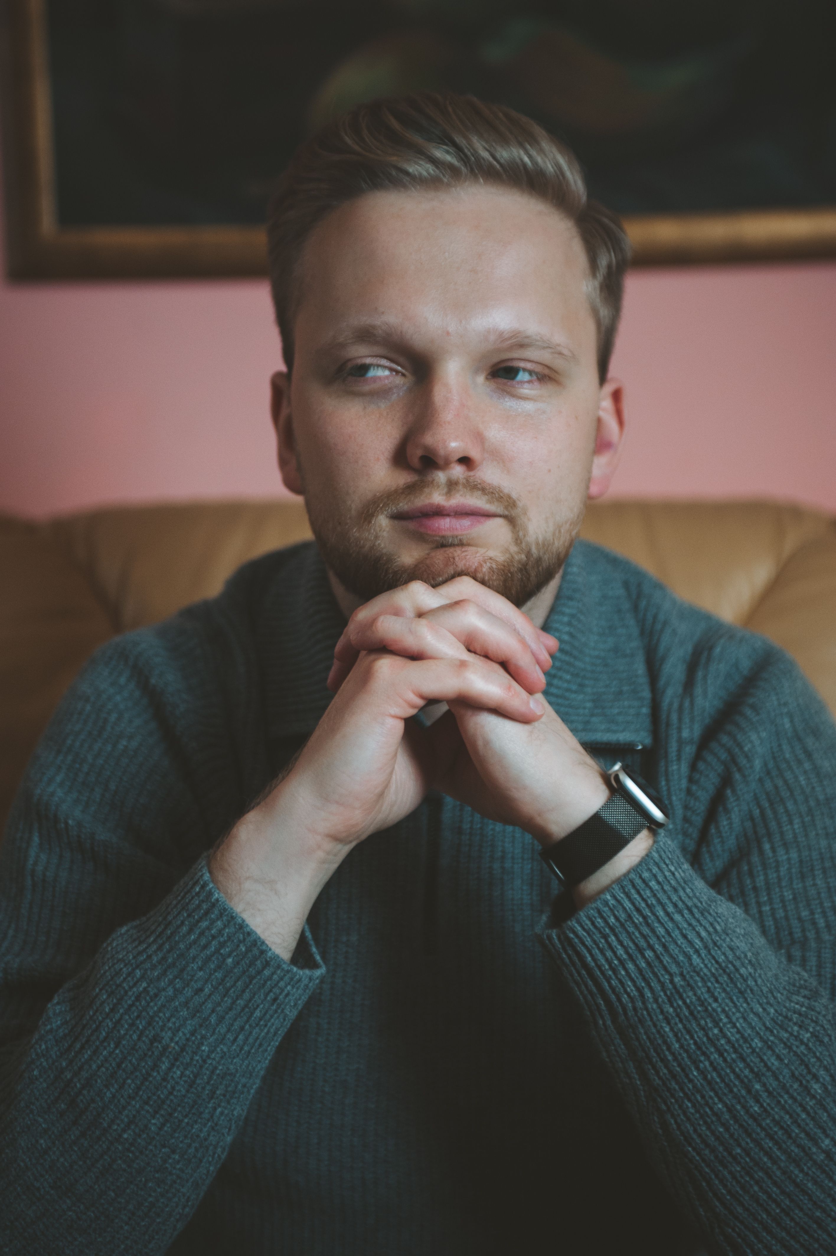 Portrait of a thoughtful man sitting with hands clasped, wearing a sweater and wristwatch indoors.