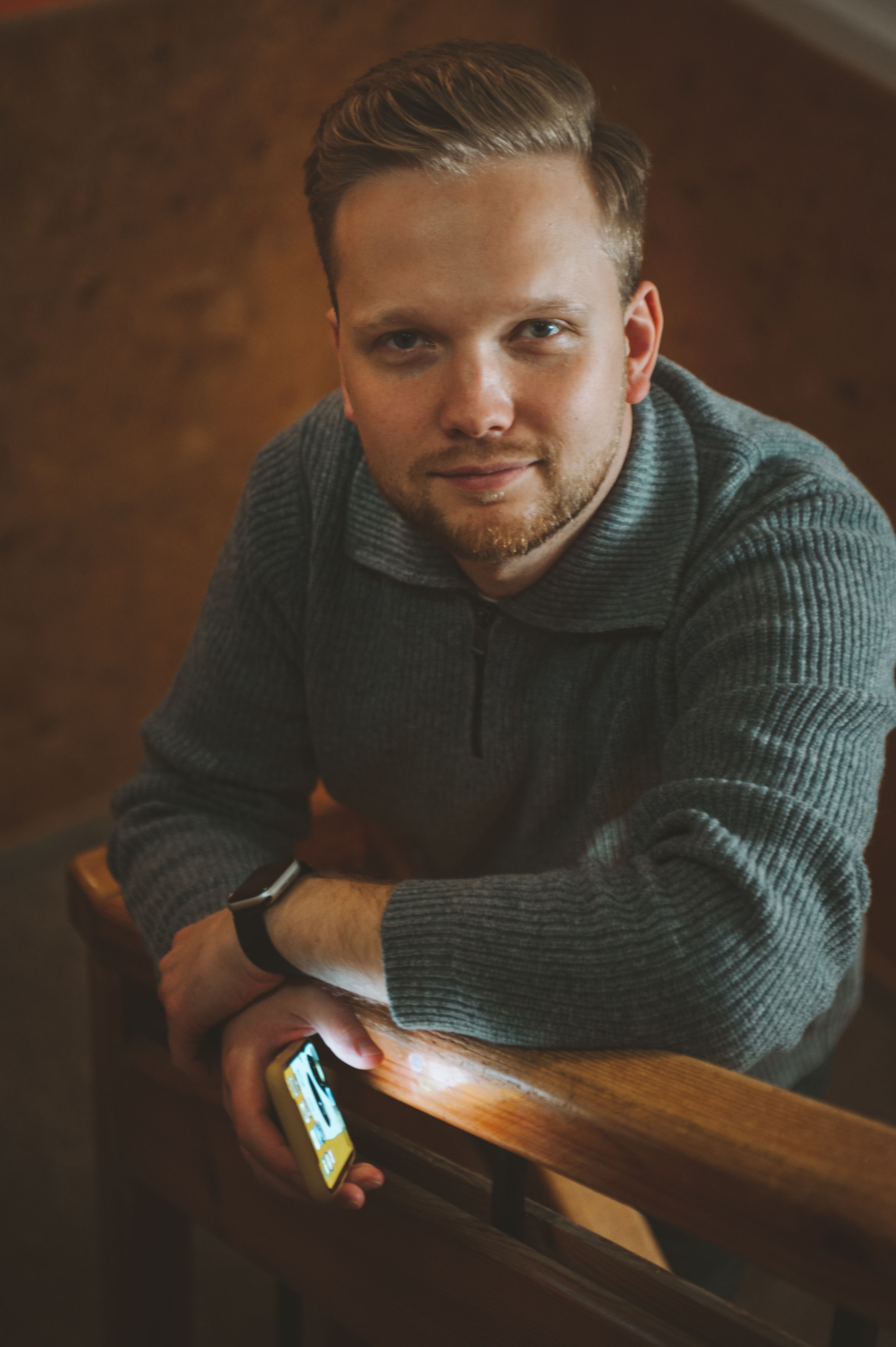 A man with short blonde hair poses casually in a cozy indoor setting.