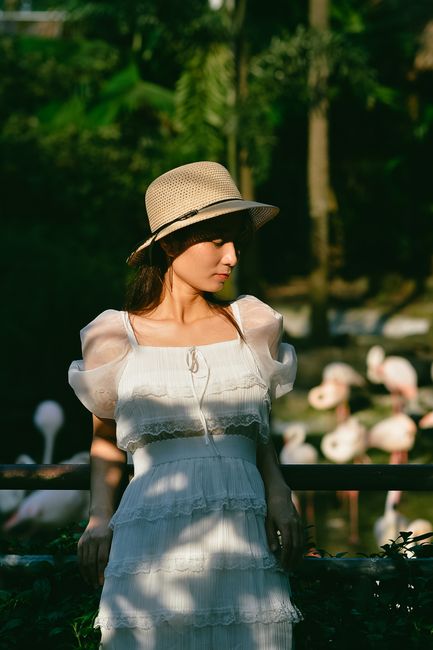 A woman in a stylish white dress and hat stands gracefully outdoors in natural light.