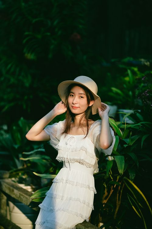 Free An Asian woman in a white dress poses gracefully in a lush garden, wearing a stylish straw hat. Stock Photo