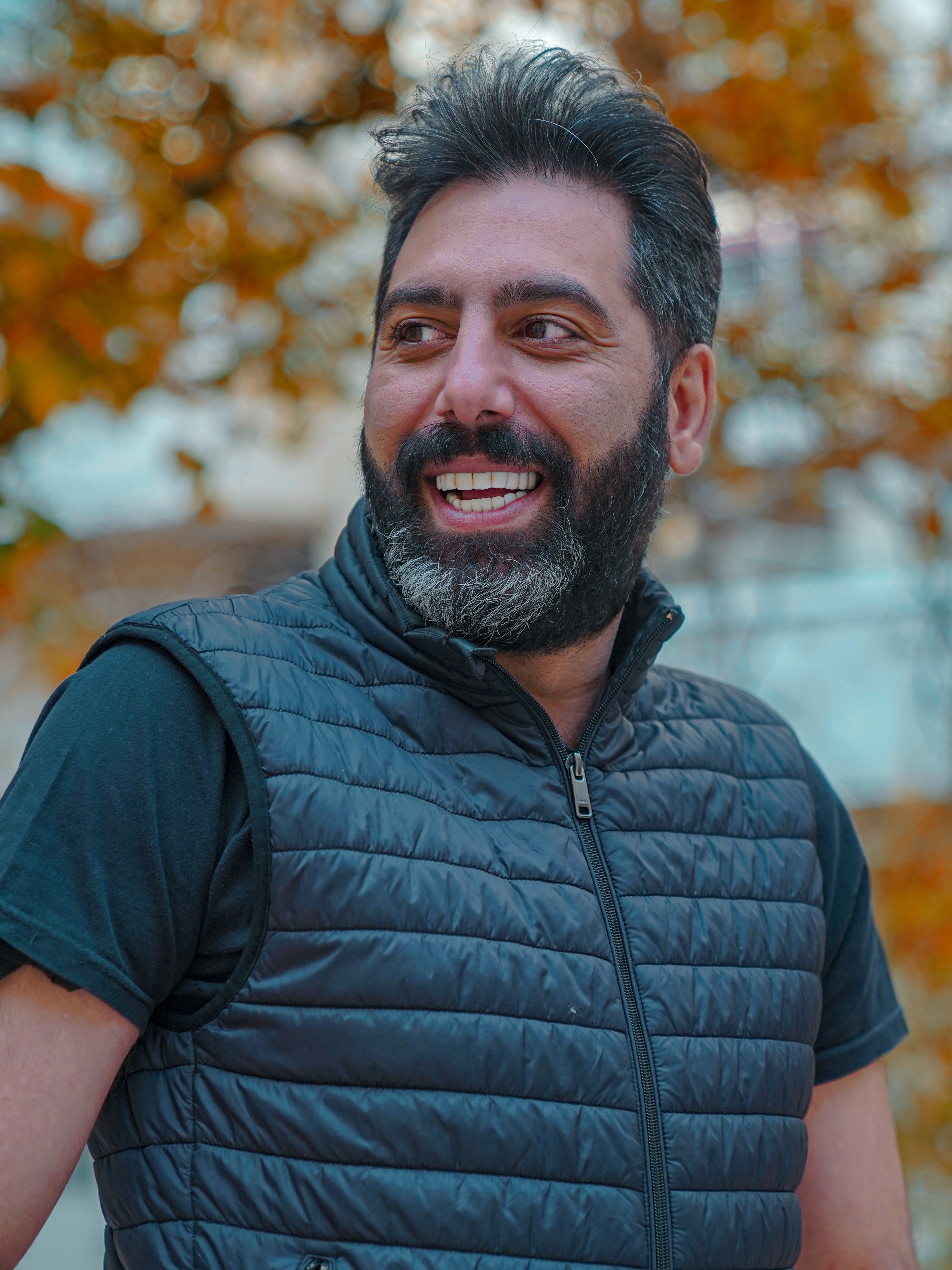 Portrait of a smiling bearded man outdoors in a quilted vest, surrounded by fall foliage.