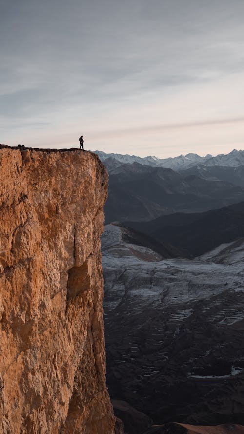 Free A lone adventurer stands on a high mountain cliff overlooking vast mountain ranges at dawn. Stock Photo