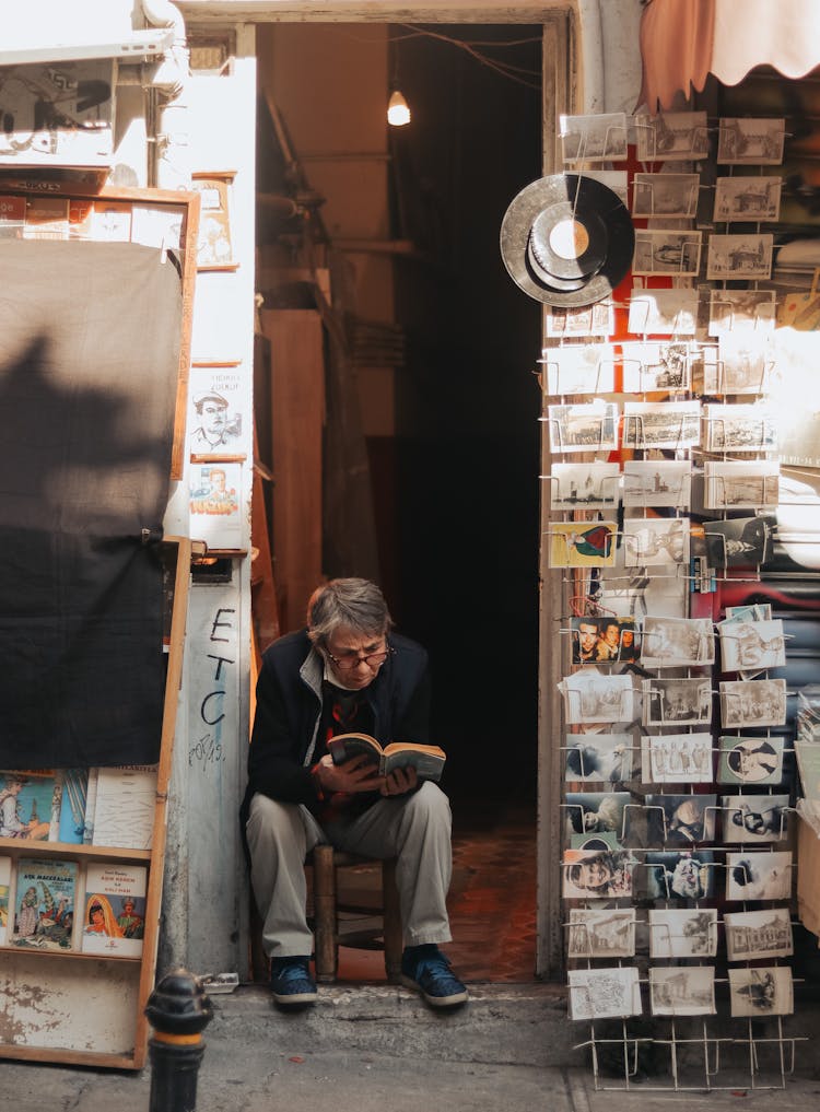 Elderly Man Sitting On Stool By Bookstore Entrance Reading Book