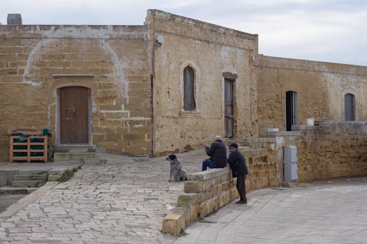 Two people and a dog sit by an ancient stone building in Gallipoli, Italy.