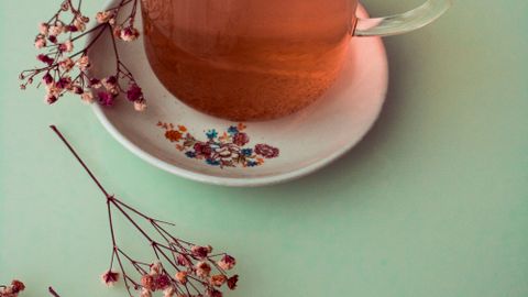 A glass of herbal tea on a floral plate with dried flowers on a pastel green background.